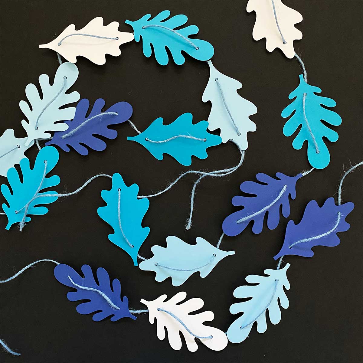 Paper leaves in shades of blue strung together as a garland on a black background for CakewalkDIY.