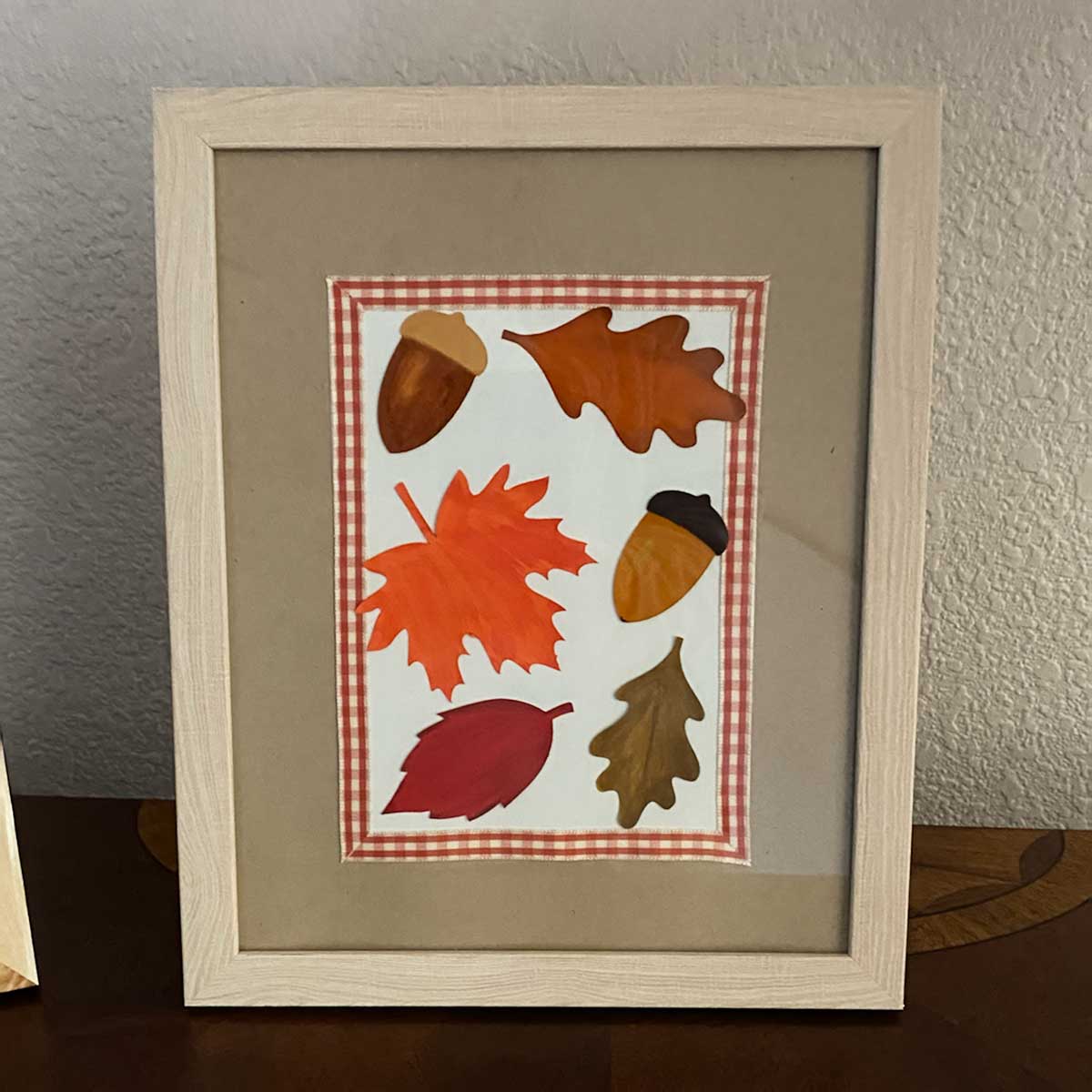 Framed artwork with autumn leaves and acorns with a checkered border to highlight a  Cakewalk DIY paper craft tutorial.