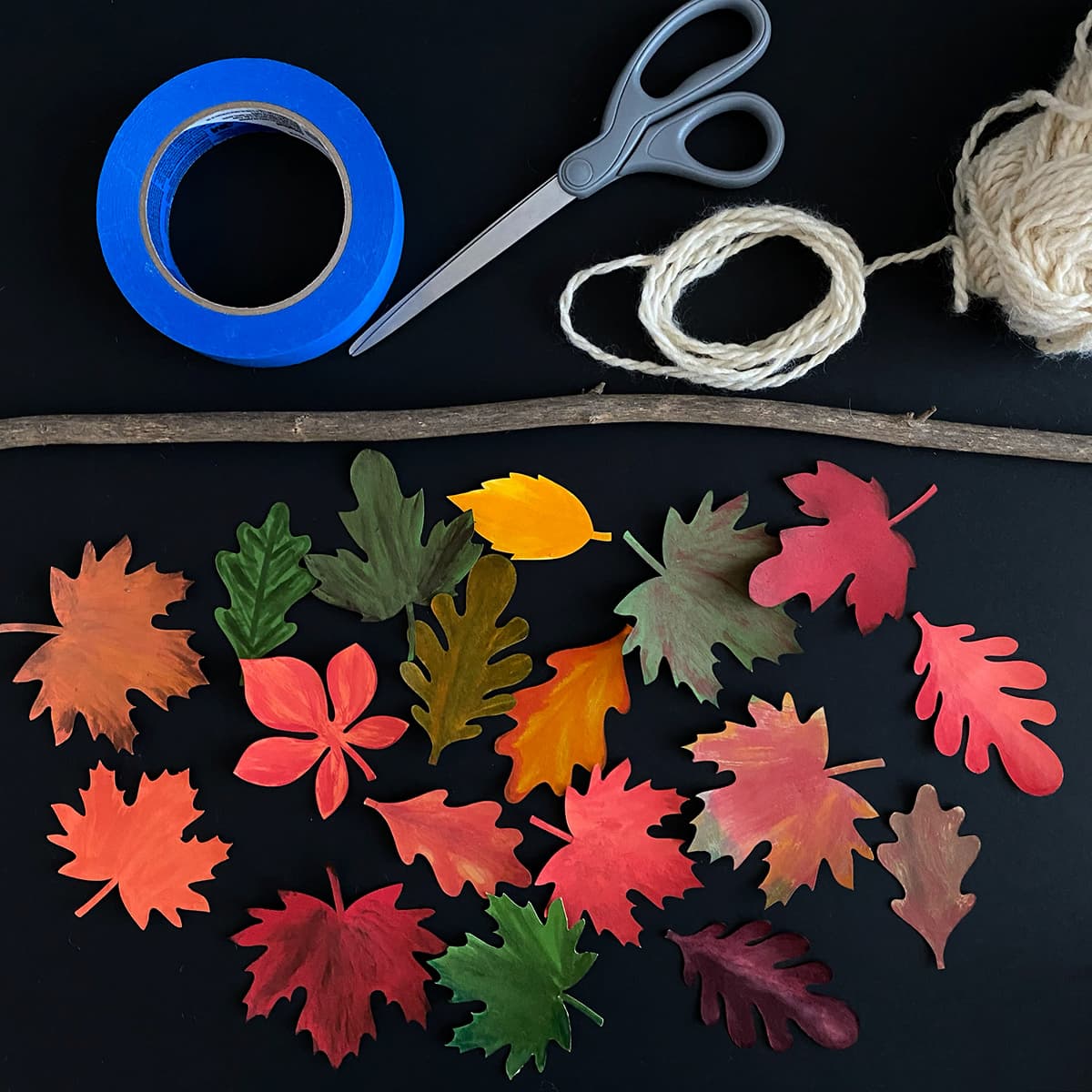 Colorful leaf-shaped cutouts with scissors, tape, twine, and a stick on a black background for a Cakewalk DIY paper Craft tutorial.