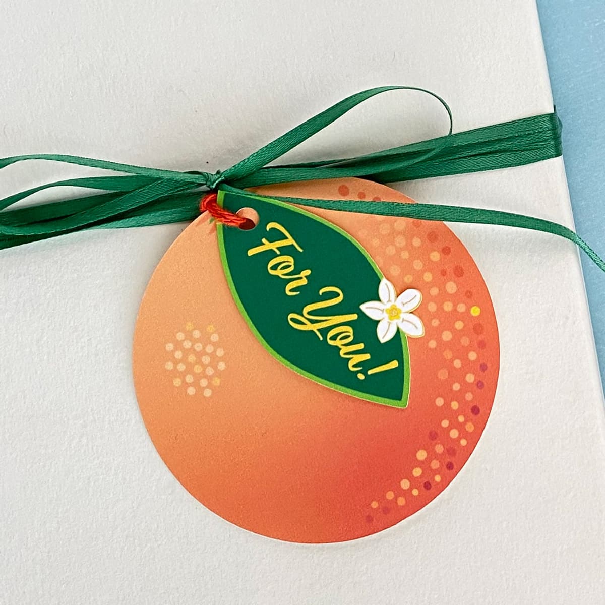 Orange Blossom gift tag with orange twine tied to a white gift box by Mozaic Studio.
