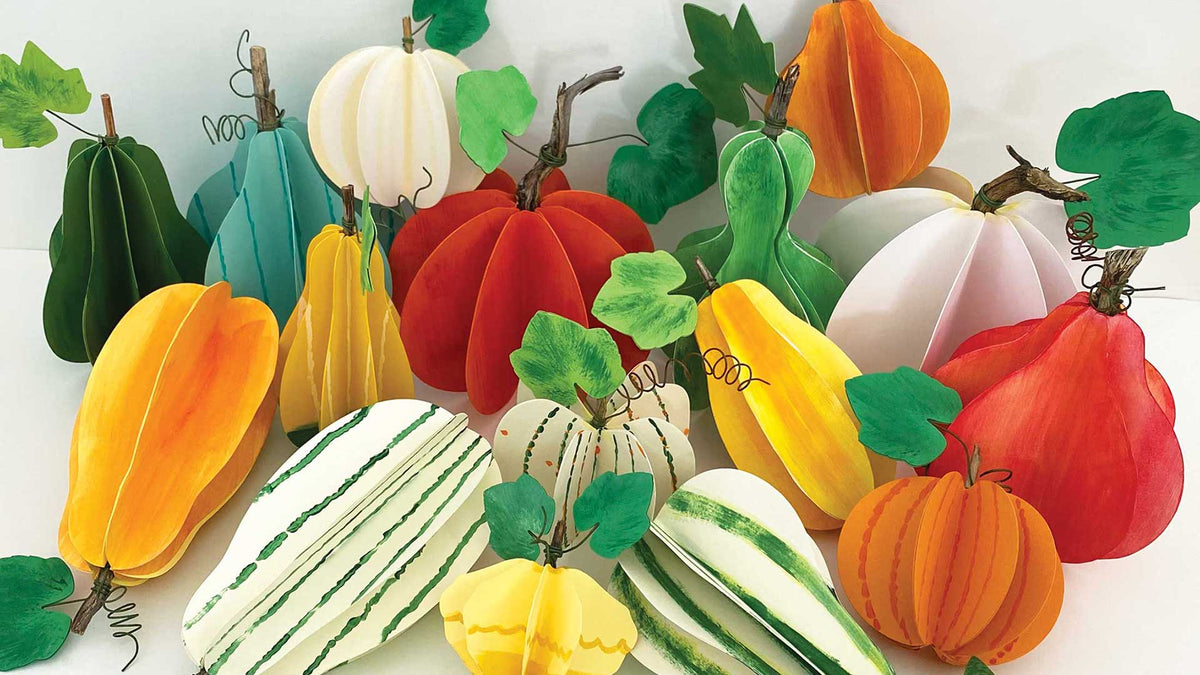 A collection of colorful, 3D papercraft pumpkins and gourds with green leaves, arranged together on a plain white background.