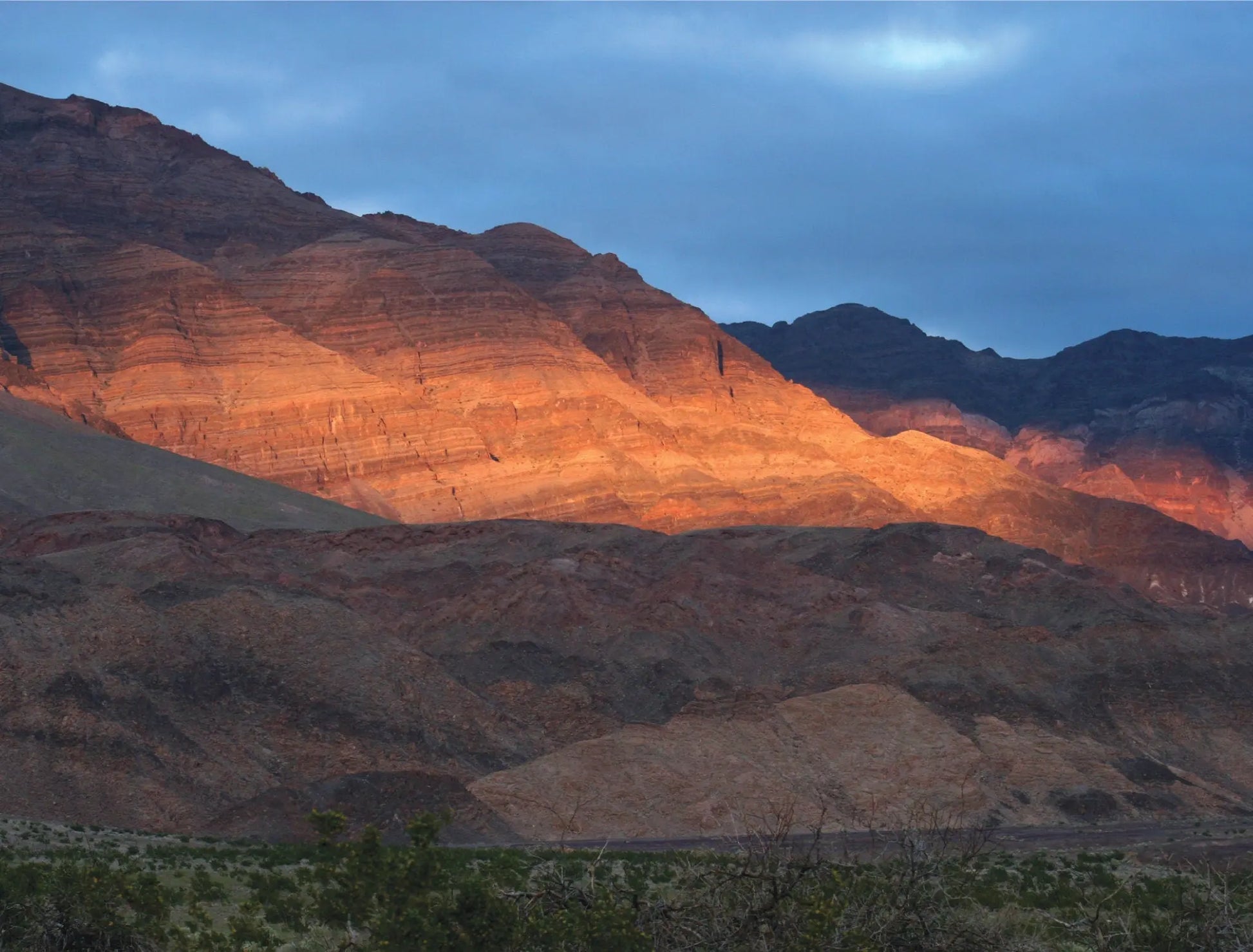 A photo titled "Winter Warmth" captures the contrasting elements of a Death Valley winter sunset.  A rugged mountain range dominates the background, its face highlighted by a band of golden sunlight that cuts through the cloudy sky.